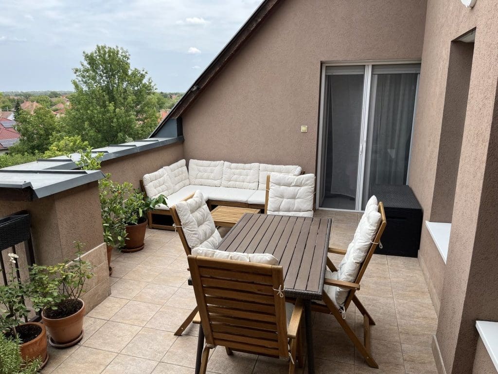 Tetőtéri terasz kerti bútorokkal, párnázott ülőgarnitúrával és növényekkel, kilátással családi házas környezetre.