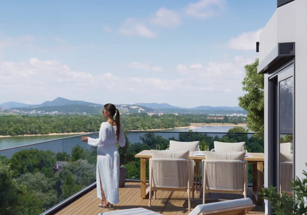 Látványterv a Waterside Residence egyik lakásának dunai panorámás kilátásáról. A képen egy tágas teraszon egy fehér ruhába öltözött fiatal nő gyönyörködik a tájban.