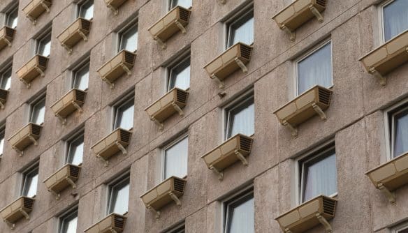 Az épület homlokzatán szabályos rácsszerkezetben elhelyezkedő ablakok láthatók, mindegyik alatt azonos típusú, bézs színű külső légkondicionáló egységgel. A szürke, kissé kopott beton- vagy vakolt felület ipari, paneles jellegre utal. Az ablakok egyformák, fehér keretesek, függönnyel vagy redőnnyel részben takartak. A homlokzat monoton ismétlődése és a klímaberendezések sora sűrűn lakott, többlakásos ház benyomását kelti.