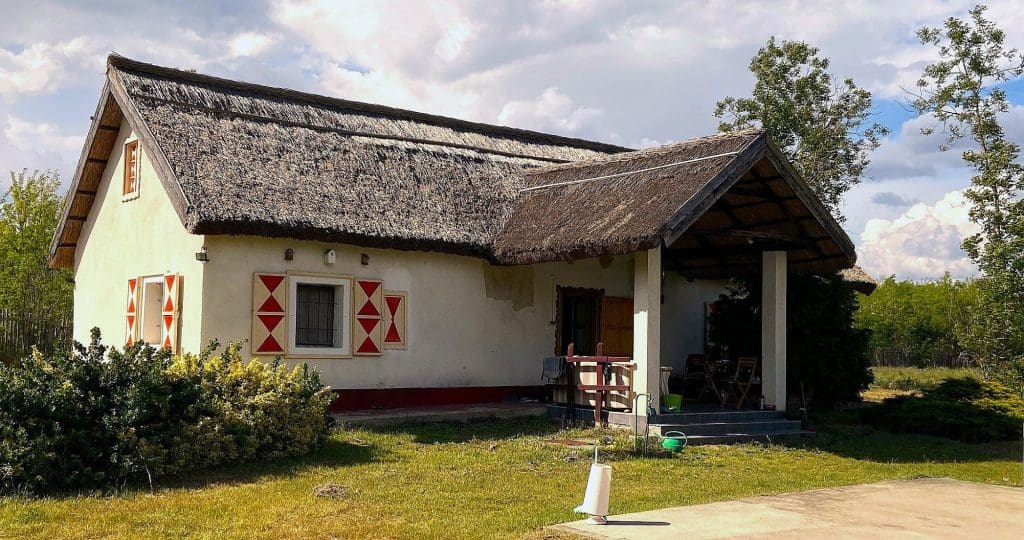 Hagyományos, nádtetős vidéki parasztház fehér falakkal és piros díszítésű zsalugáterekkel, előtte tornác és füves udvar.