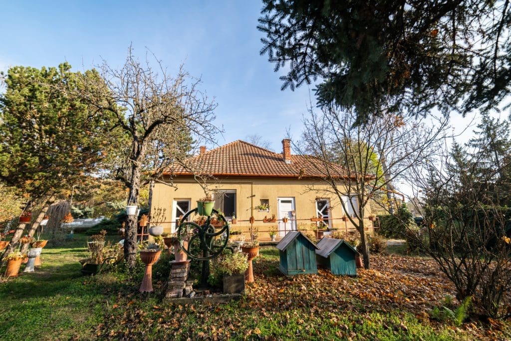 Sárga falú családi ház nagy, parkosított kerttel, fákkal, virágcserepekkel és kis díszkutakkal.