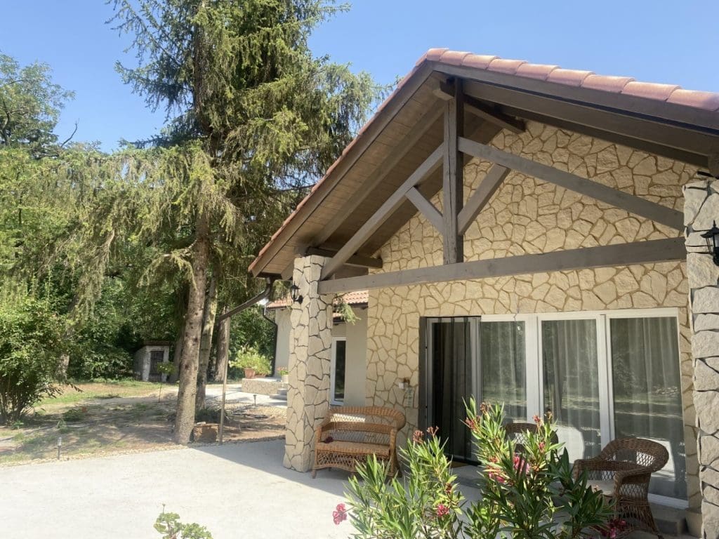 Terméskő burkolatú, modern családi ház fedett terasszal, kerti bútorokkal és fákkal körülvéve.