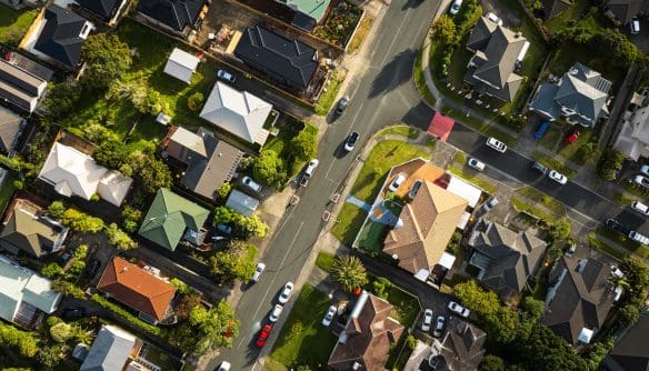 Légi felvétel egy kertvárosi lakóövezetről, ahol családi házak sorakoznak rendezett telkeken. Az utcák sakktáblaszerűen találkoznak egy kereszteződésben, több autó halad és parkol az utak mentén. A házak különböző színű tetőkkel és zöld kertekkel rendelkeznek, a környezetet fák és gondozott udvarok teszik barátságossá.