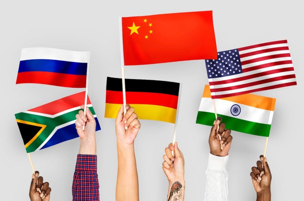 Emberi kezek különböző országok zászlóit tartják fel egy fehér háttér előtt. A kép a külföldi ingatlanvásárlókról szóló cikket illusztrálja.