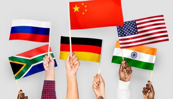 Emberi kezek különböző országok zászlóit tartják fel egy fehér háttér előtt. A kép a külföldi ingatlanvásárlókról szóló cikket illusztrálja.