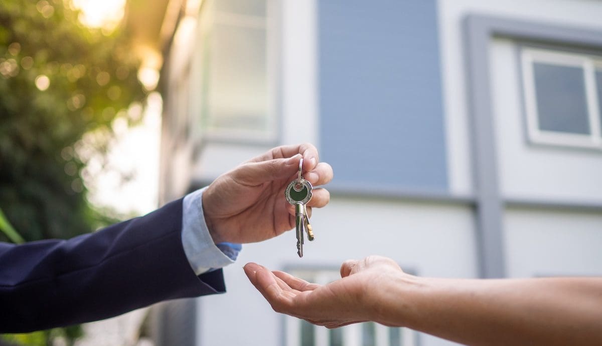 Ingatlanvásárlók átveszik a ház kulcsait az eladótól. A képen két kéz látható, az egyik egy kulcsot nyújt a másiknak. Mögöttük egy kék családi ház részlete elmosódva.