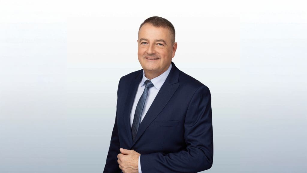 Egy középkorú férfi öltönyben és nyakkendőben, semleges világos háttér előtt állva mosolyog a kamerába. Megjelenése elegáns és üzleties, portré jellegű fotó, amely professzionális vagy vállalati környezetre utal.