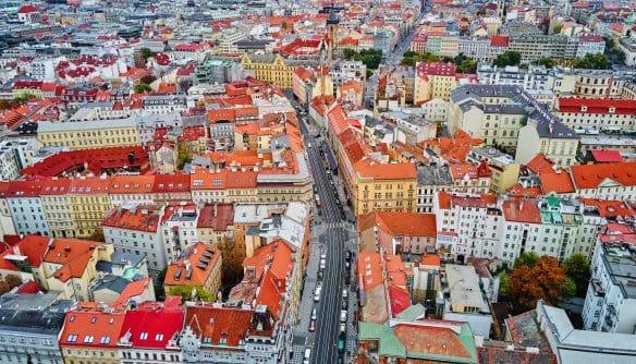 Légi felvétel egy sűrűn beépített történelmi belvárosról, ahol szorosan egymás mellett álló, többemeletes házak és vörös cseréptetők uralják a látképet. A városszövetet egy hosszú, egyenes út szeli ketté, az út mentén autók sorakoznak és haladnak. A kép sokszínű homlokzatú, klasszikus európai városi épületeket mutat, a háttérben templomtornyokkal és nagyobb középületekkel. A felvétel a város sűrű, történelmi jellegét és egységes tetővilágát hangsúlyozza.