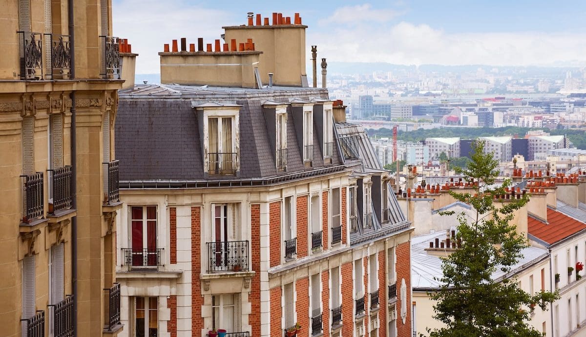 Városi utcakép, többemeletes, régi építésű lakóházakkal, jellegzetes tetőkkel és kéményekkel. Az előtérben egy díszes homlokzatú épület látható erkélyekkel, a háttérben sűrűn beépített városrész és távolabbi városi panoráma.