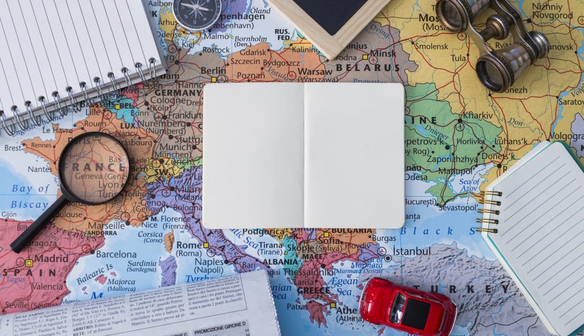 Európa térképén elhelyezett utazási kellékek – nagyító, iránytű, távcső és jegyzetfüzet – egy nyitott üres füzet körül, amely az utazástervezés és felfedezés hangulatát idézi.