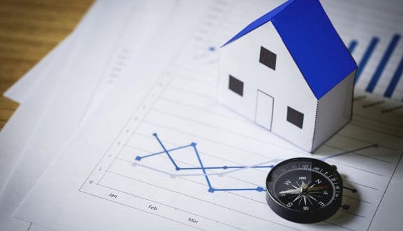 Makett ház grafikonokkal ábrázolt pénzügyi kimutatásokon, mellette iránytű; az ingatlanpiaci elemzés, befektetési döntések és trendkövetés vizuális megjelenítése.