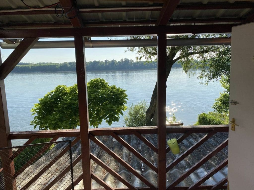 Fedett teraszról nyíló kilátás egy nyugodt folyóra, előtérben fa korlát és tetőszerkezet, körülötte zöld növényzet; a jelenet vízparti ingatlan csendes, természetközeli környezetét mutatja.