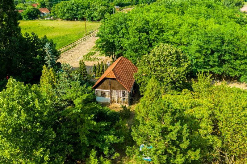 Légifelvétel egy kis vidéki házról vörös tetővel, sűrű zöld növényzettel és fákkal körülvéve; a háttérben mezők és egy földút látható, amely nyugodt, természetközeli környezetet sugall.