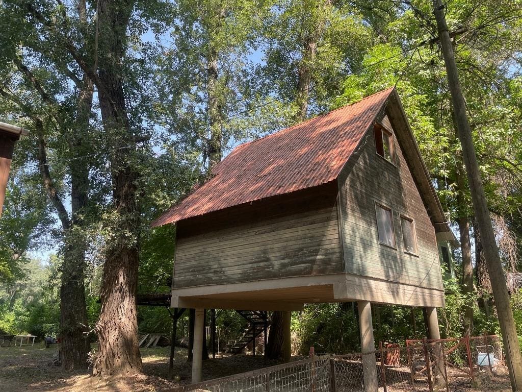 Egy kis, fa borítású nyaralóház látható, amely oszlopokra emelve áll egy fás, árnyékos területen; a ház vöröses tetővel rendelkezik, körülötte magas fák és természetközeli környezet található.