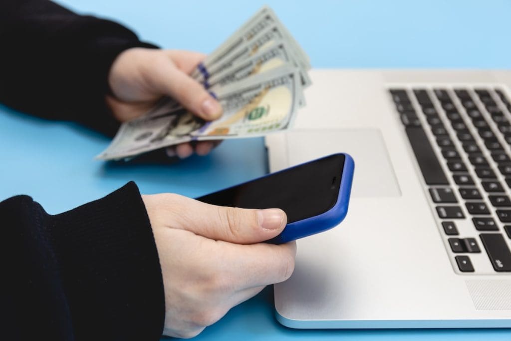 Egy személy okostelefont tart a kezében laptop mellett, miközben a másik kezében amerikai dollár bankjegyeket fog; a kép az online fizetési kérelem, digitális tranzakciók vagy pénzügyi ügyintézés témáját szemlélteti.
