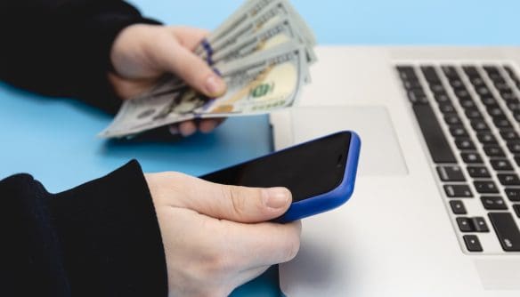 Egy személy okostelefont tart a kezében laptop mellett, miközben a másik kezében amerikai dollár bankjegyeket fog; a kép az online fizetési kérelem, digitális tranzakciók vagy pénzügyi ügyintézés témáját szemlélteti.
