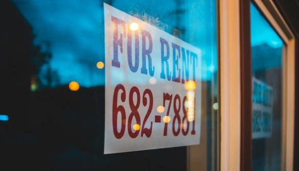 „For Rent” feliratú kiadó lakást hirdető tábla látható egy kirakatüvegen esti fényekkel és elmosódott utcai háttérrel.