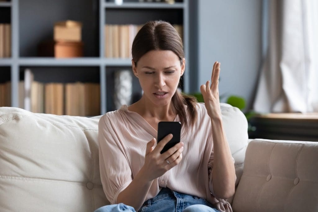 Egy világos nappaliban, kanapén ülő nő bosszúsan nézi a mobiltelefonját, miközben egyik kezét értetlenkedve a magasba emeli.