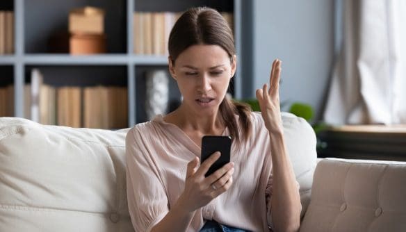Egy világos nappaliban, kanapén ülő nő bosszúsan nézi a mobiltelefonját, miközben egyik kezét értetlenkedve a magasba emeli.