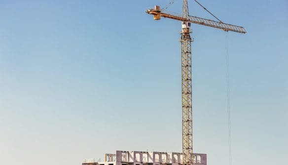 Magas, sárga építési daru magasodik egy épülő, szerkezetkész többemeletes társasház mellett, tiszta, kék égbolt előtt. A kép tökéletesen szemlélteti az új építésű lakások piacát, a lakópark-fejlesztéseket, az építőipari beruházásokat és a lakáskínálat bővülését.