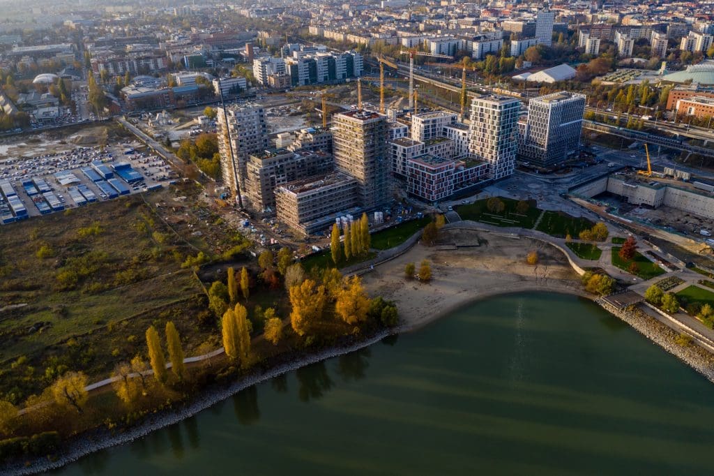 Magasból készült légi fotó egy vízparti, modern új építésű lakóparkról és irodanegyedről, építési darukkal, parkosított öböllel és a vízpartot szegélyező, sárguló őszi fákkal. A látványos felvétel jól szemlélteti a nagyszabású városfejlesztési beruházásokat, a vízparti prémium ingatlanok piacát és a modern lakóparki projektfejlesztéseket.