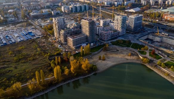 Magasból készült légi fotó egy vízparti, modern új építésű lakóparkról és irodanegyedről, építési darukkal, parkosított öböllel és a vízpartot szegélyező, sárguló őszi fákkal. A látványos felvétel jól szemlélteti a nagyszabású városfejlesztési beruházásokat, a vízparti prémium ingatlanok piacát és a modern lakóparki projektfejlesztéseket.