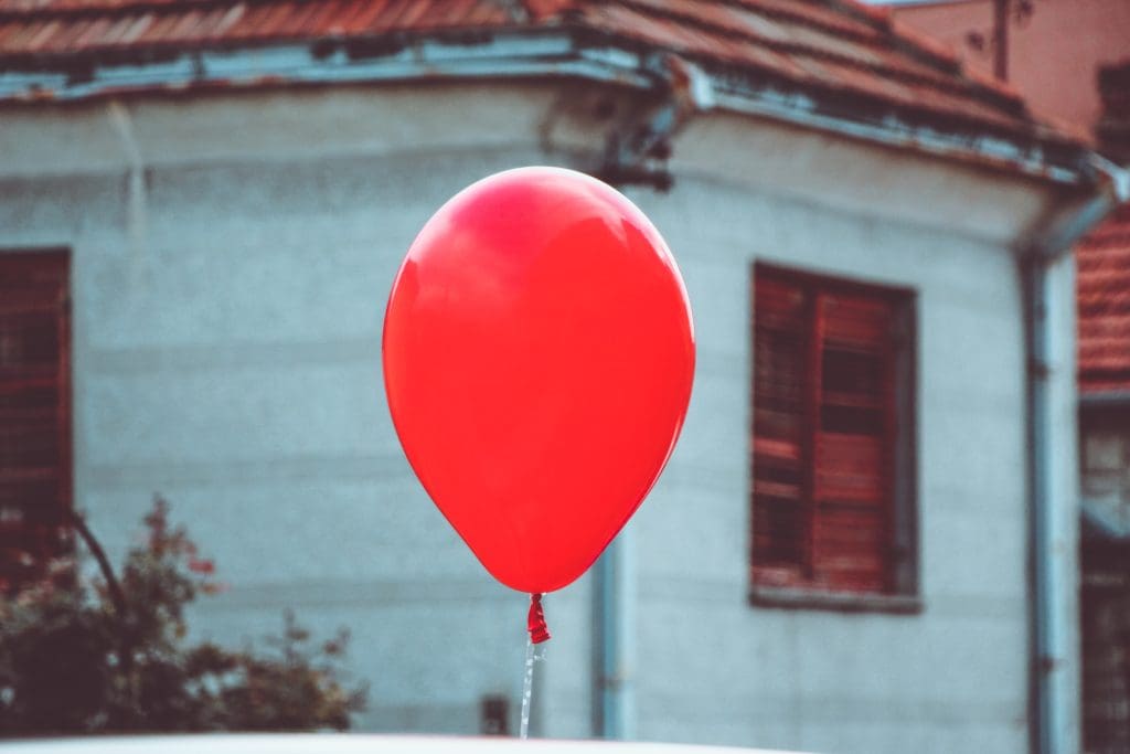 Közeli felvétel egy levegőben lebegő, élénkpiros lufiról, a háttérben egy elmosódott, régi cseréptetős ház homlokzatával. A szimbolikus kép tökéletesen illusztrálja az ingatlanpiaci buborék jelenségét, a lakásárak túlfűtöttségét, a túlárazottság kockázatait vagy a piaci lufi kidurranásának veszélyét.