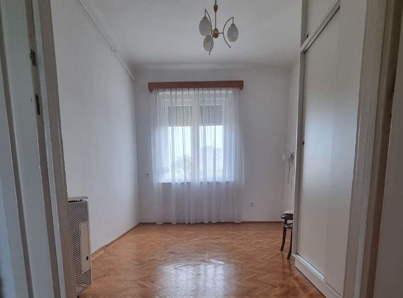 Ajtókeretből fényképezett, világos és üres szoba klasszikus halszálka mintás fa parkettával, fehér falakkal és a helyiség végén lévő, fehér függönyös ablakkal. A szobában egy gázkonvektor, egy beépített tolóajtós szekrény és egy hármas ágú csillár látható. A kép kiválóan illusztrálja a bútorozatlan, költözhető állapotú eladó vagy kiadó lakásokat és a tradicionális polgári vagy téglalakások kínálatát.