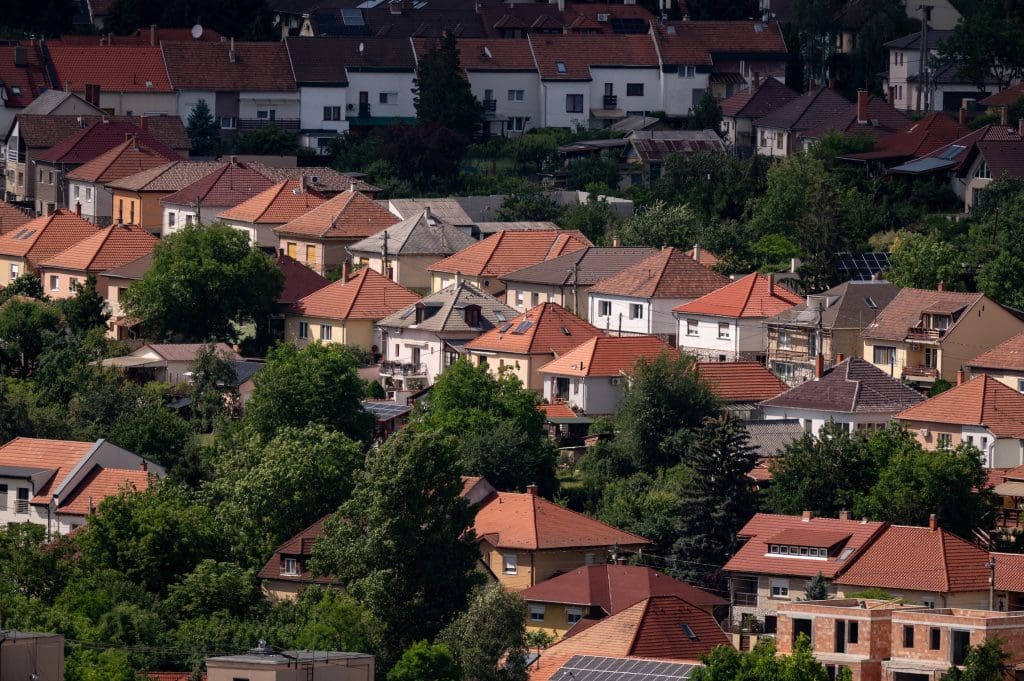 Magasból készült fotó egy sűrűn beépített, domboldali lakóövezetről, piros cseréptetős családi házakkal és kertes ingatlanokkal, zöld fák ölelésében. A kép tökéletesen illusztrálja a kertvárosi ingatlanpiacot, a családi házak kínálatát és a zöldövezeti lakhatási lehetőségeket.