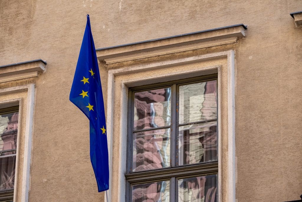 Az Európai Unió kék, sárga csillagos zászlaja függőlegesen kitűzve egy klasszikus, bézs homlokzatú épület ablaka mellett. A fotó tökéletesen illusztrálja az európai uniós szabályozásokat, az EU-s támogatásokat, a nemzetközi gazdasági folyamatokat és a hazai ingatlanpiacot érintő uniós direktívákat.