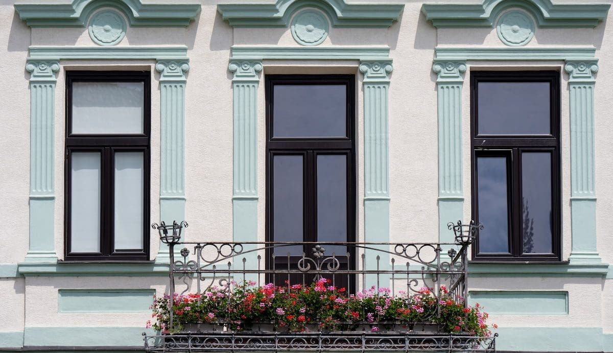 Díszes, történelmi jellegű épület homlokzata három magas ablakkal, pasztellzöld stukkódíszekkel és kovácsoltvas erkéllyel, amelyet virágok díszítenek.
