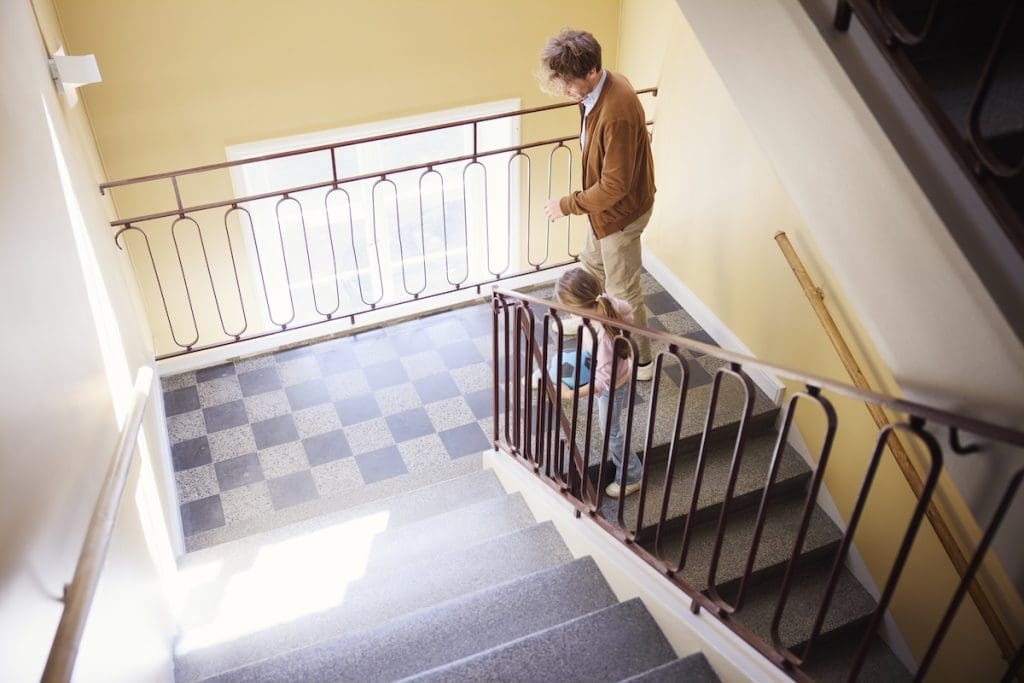 Lakóépület lépcsőháza. Egy apuka és kislánya felülről láthatóak, ahogy sétálnak fel a lépcsőn.