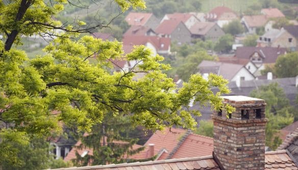 Magasból készült fotó egy cseréptetős ház tégla kéményéről, amely mögött a fák lombjai között egy domboldali kistelepülés piros tetős házai terülnek el. A hangulatos, vidékies kép kiválóan illusztrálja a kistelepülési és falusi ingatlanpiacot, a vidékre költözést, a családi házak kínálatát, valamint az otthoni fűtési megoldásokat.