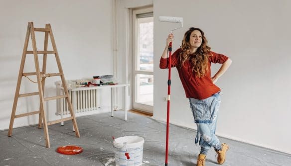 Felújítás közben festőhengerrel pózoló nő egy világos, üres szobában, festékesvödörrel, létrával és felújítási eszközökkel a háttérben.